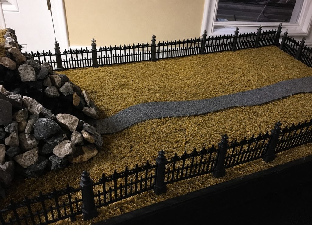 Halloween village landscape with a winding road, moss ground cover, rock wall, and black fencing.