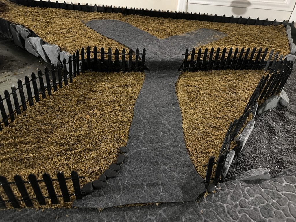 Halloween village roads and pathways made from dark textured material, bordered by fences, rocks, and ground cover.