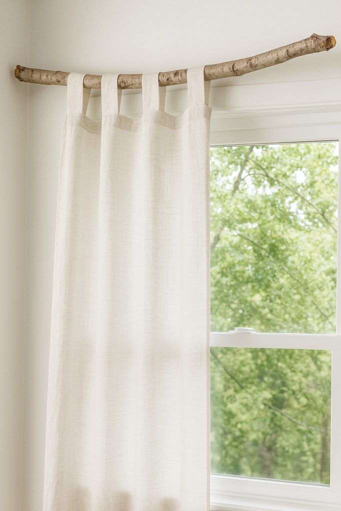 White tab-top curtains hanging from a natural tree branch above a window with green foliage outside — a rustic DIY curtain rod idea.