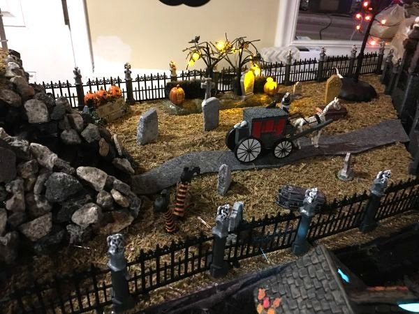 Halloween village cemetery area with fences, rock landscaping, a winding path, gravestones, lights, and a horse-drawn carriage.